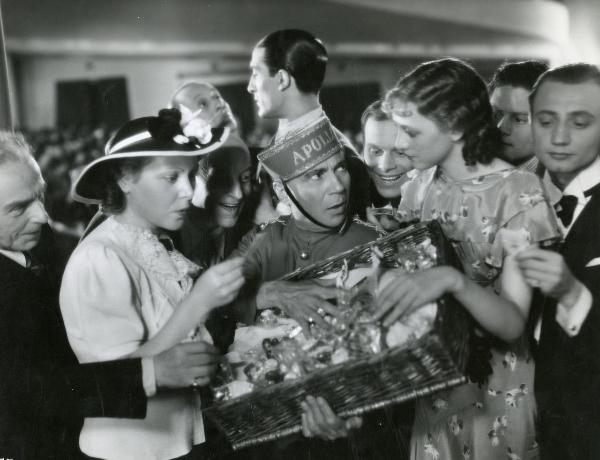 Scena del film "Il feroce Saladino" - Bonnard, Mario, 1937 - Al centro, un attore non identificato osserva un'attrice non identificata alla sua sinistra che appoggia una mano sulla merce. Tutt'intorno sono presenti attori non identificati.
