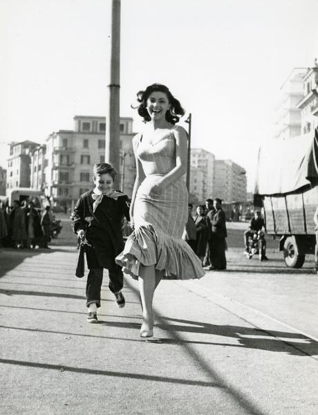 Scena del film "Il ferroviere" - Germi, Pietro, 1956 - Sylva Koscina, a destra, e Edoardo Nevola, a sinistra, in divisa scolastica, corrono sorridenti lungo un marciapiede osservati da attori non identificati.
