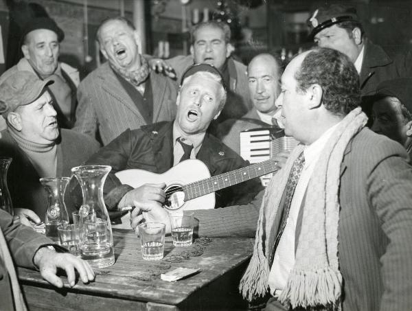 Scena del film "Il ferroviere" - Germi, Pietro, 1956 - Intono a un tavolo: Pietro Germi, al centro, suona una chitarra e canta insieme ad altri attori non identificati che lo circondano.
