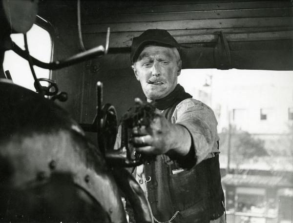 Scena del film "Il ferroviere" - Germi, Pietro, 1956 - Mezza figura di Pietro Germi che, nella cabina motore di un treno, con un sigaro in bocca, armeggia con i comandi e rivolge lo sguardo in basso a destra.

