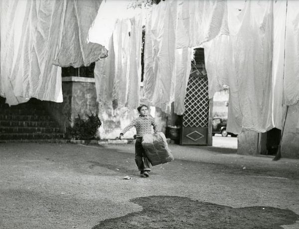 Scena del film "Il ferroviere" - Germi, Pietro, 1956 - In un cortile, tra panni stesi, il giovane Edoardo Nevola cammina verso l'obbiettivo, trasportando un enorme scatola di cartone chiusa.
