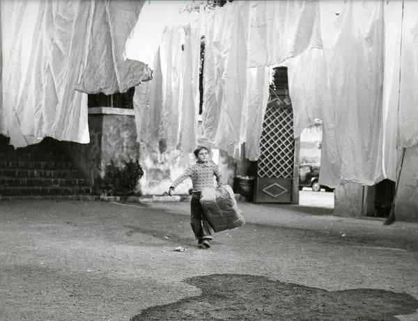 Scena del film "Il ferroviere" - Germi, Pietro, 1956 - In un cortile, tra panni stesi, il giovane Edoardo Nevola cammina verso l'obbiettivo, trasportando un enorme scatola di cartone chiusa.
