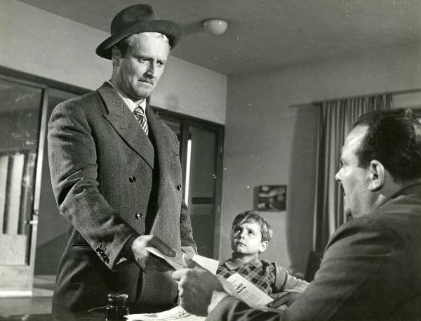 Scena del film "Il ferroviere" - Germi, Pietro, 1956 - Pietro Germi, a sinistra, conversa con un attore non identificato, seduto a destra. Entrambi tengono tra le mani un foglio di giornale. Intanto, al centro, Edoardo Nevola, osserva Pietro Germi.
