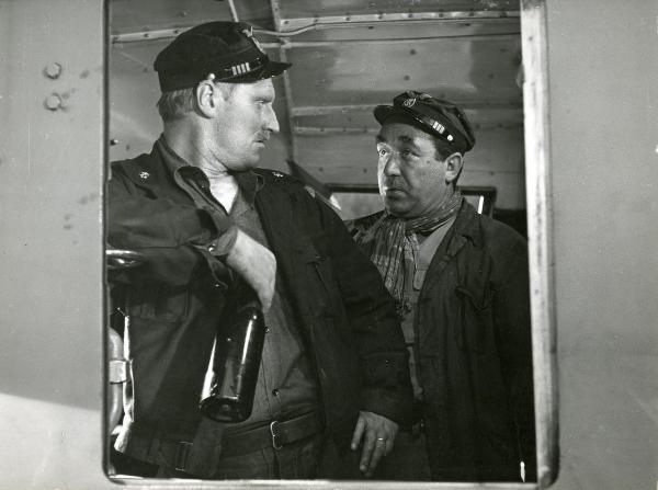 Scena del film "Il ferroviere" - Germi, Pietro, 1956 - Dal finestrino di un treno si vedono Pietro Germi, a sinistra con una bottiglia di vino in mano, e Saro Urzì, a destra, mentre si osservano.
