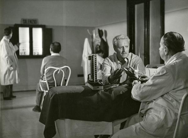 Scena del film "Il ferroviere" - Germi, Pietro, 1956 - Pietro Germi, a destra, discute con un attore non identificato di spalle. Dietro, un attore non identificato, vestito da medico, effettua un'esame della vista a un altro attore non identificato.