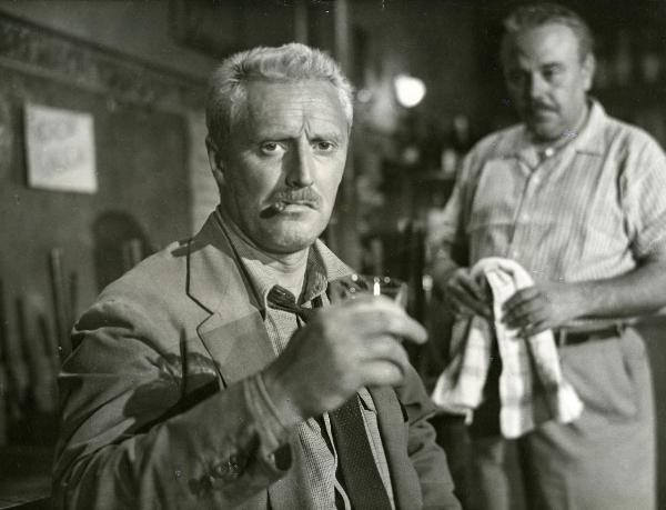 Scena del film "Il ferroviere" - Germi, Pietro, 1956 - Primo piano di Pietro Germi che, con un sigaro in bocca e un bicchiere in mano, rivolge lo sguardo verso l'obbiettivo. Dietro, un attore non identificato, con un canovaccio in mano, lo osserva.