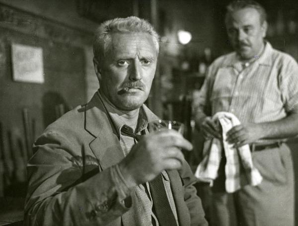 Scena del film "Il ferroviere" - Germi, Pietro, 1956 - Primo piano di Pietro Germi che, con un sigaro in bocca e un bicchiere in mano, rivolge lo sguardo verso l'obbiettivo. Dietro, un attore non identificato, con un canovaccio in mano, lo osserva.