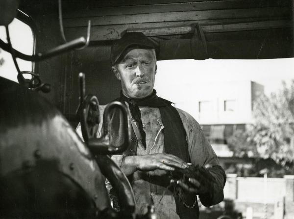 Scena del film "Il ferroviere" - Germi, Pietro, 1956 - In una cucina: Mezza figura di Pietro Germi che, con un sigaro in bocca, nella cabina motore di un treno, si pulisce le mani con una pezza sporca.
