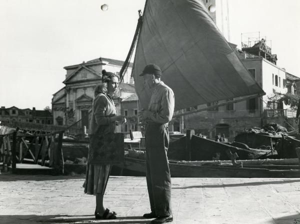 Scena del film "Fiamme sulla laguna" - Scotese, Giuseppe Maria, 1951 - Con alle spalle un paesaggio veneziano, Lea Padovani, a sinistra con una borsa al braccio, discute con Lauro Gazzolo, a destra con le mani giunte.
