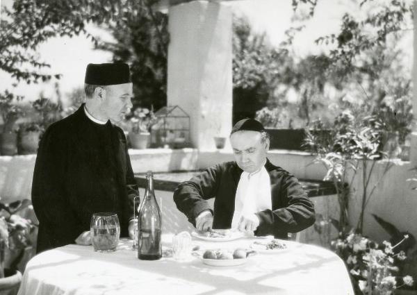 Scena del film "Fiat voluntas Dei" - Palermi, Amleto, 1935 - Un attore non identificato in piedi a sinistra osserva Angelo Musco, a destra seduto a tavola mentre mangia. Entrambi indossano abiti talari.
