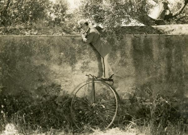 Scena del film "Figaro e la sua gran giornata" - Camerini, Mario, 1931 - Maurizio D'Ancora, con un cappello a bombetta in testa, in piedi sul sellino di un velocipede, si appoggia a un muro mentre guarda a destra.
