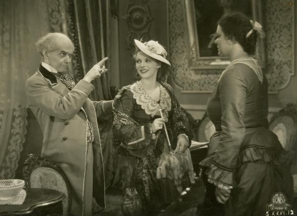 Scena del film "Figaro e la sua gran giornata" - Camerini, Mario, 1931 - Gloria Leda, con un ombrello in mano e un cappello in testa, e un'attrice non identificata, a destra, guardano verso Gianfranco Giachetti, a sinistra che indica davanti a sé.
