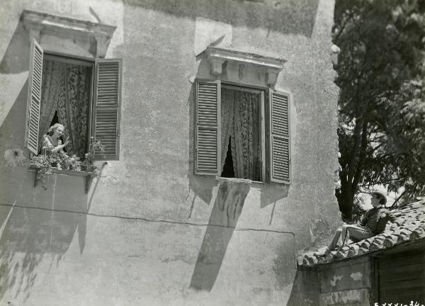 Scena del film "Figaro e la sua gran giornata" - Camerini, Mario, 1931 - Gloria Leda, a sinistra affacciata a una finestra, saluta in direzione di Maurizio D'Ancora, steso sul tetto di un edificio, a destra con un cappello a bombetta.
