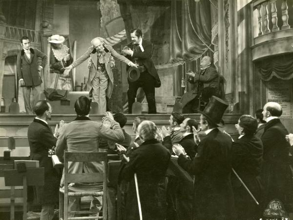 Scena del film "Figaro e la sua gran giornata" - Camerini, Mario, 1931 - Gianfranco Giachetti fa un inchino di ringraziamento. Gloria Leda, gli si avvicina e Maurizio D'Ancora, a sinistra lo osserva. Attorno, attori non identificati lo applaudono.