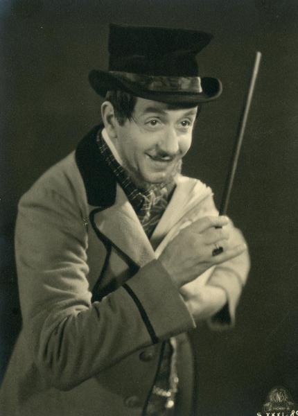 Scena del film "Figaro e la sua gran giornata" - Camerini, Mario, 1931 - Primo piano di un attore non identificato che, tenendo sollevata con la mano destra una bacchetta e tenendo un plico di fogli con la mano sinistra, guarda sorridente a destra.
