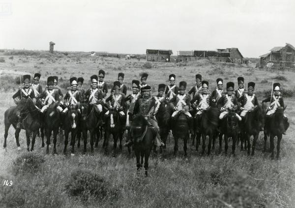 Scena del film "La figlia del capitano" - Camerini, Mario, 1947 - In un paesaggio di campagna con degli edifici in legno sullo sfondo, una schiera di attori non identificati a cavallo, in abiti militari, rivolgono lo sguardo verso l'obbiettivo.
