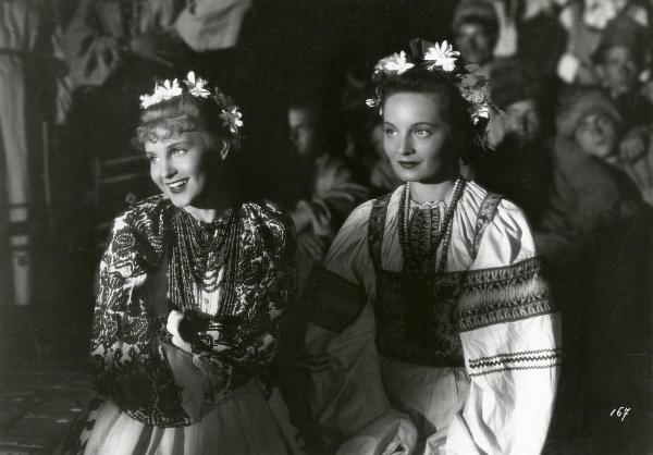 Scena del film "La figlia del capitano" - Camerini, Mario, 1947 - Mezze figure sorridenti di Irasema Dilian, a sinistra, e un'attrice non identificata a destra, in abiti tradizionali. In secondo piano, attori non identificati.

