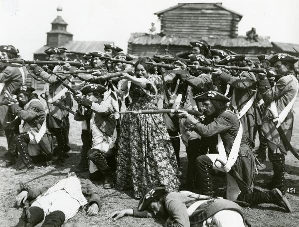 Scena del film "La figlia del capitano" - Camerini, Mario, 1947 - Olga Solbelli, al centro, imbraccia un fucile e mira verso sinistra. In primo piano, due attori non identificati sono stesi a terra, uno prono e uno supino, morti.
