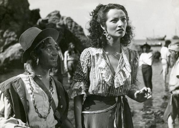 Scena del film "La figlia del corsaro verde" - Guazzoni, Enrico, 1940 - Polidor, a destra con un cappello in capo, osserva sorridente Doris Duranti, a destra con la mano sinistra alzata alla vita, che rivolge lo sguardo dritto davanti a sé.
