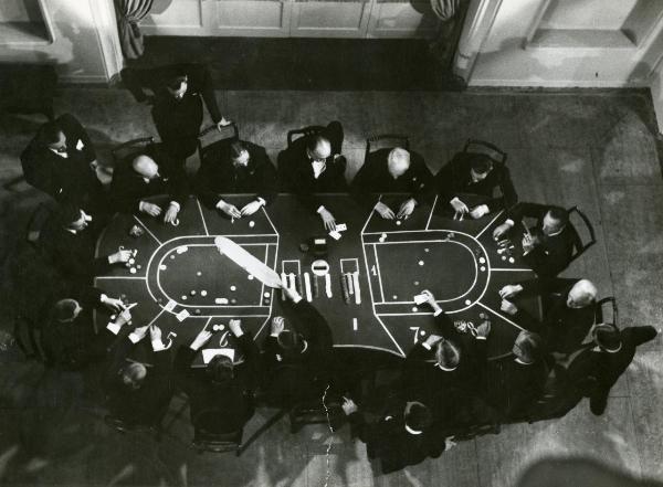 Scena del film "I figli del Marchese Lucera" - Palermi, Amleto, 1938 - Una vista dell'alto di un tavolo da gioco, al cui perimetro sono seduti attori non identificati che giocano a carte e attori in piedi che osservano la scena.
