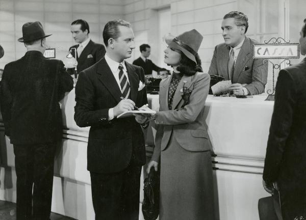 Scena del film "I figli del Marchese Lucera" - Palermi, Amleto, 1938 - Filippo Scelzo, al centro, reggendo un blocco e una penna tiene la mano di un'attrice non identificata che lo guarda.