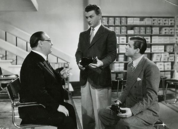 Scena del film "I figli del Marchese Lucera" - Palermi, Amleto, 1938 - Gino Cervi, tiene una scarpa in mano e conversa con un attore non identificato che solleva la mano sinistra. A destra, un attore non identificato, li osserva.
