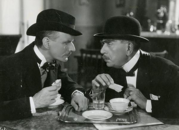 Scena del film "I figli del Marchese Lucera" - Palermi, Amleto, 1938 - Armando Falconi, con un cappello a bombetta e con un biscotto in mano, e Sergio Tofano, con un cappello in testa e una brioches in mano, si osservano con intesa.