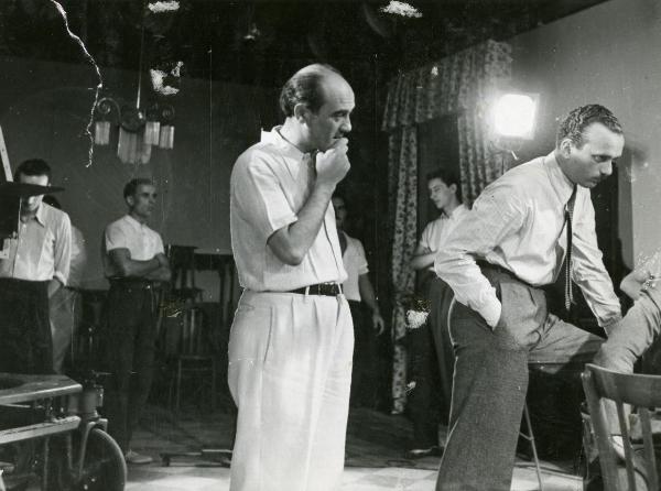 Sul set del film "I figli del Marchese Lucera" - Palermi, Amleto, 1938 - Il regista Amleto Palermi, si porta una mano al mento e guarda a destra verso un operatore non identificato che gli dà le spalle. Intorno, altri operatori non identificati.
