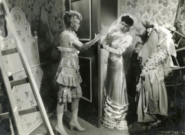 Scena del film "Finalmente soli" - Gentilomo, Giacomo, 1942 - In una piccola stanza, a sinistra, di fronte a una catasta di sedie coperte di abiti, Maria Mercader, in lingerie, tiene aperta una porta e osserva Anna Magnani, in abiti da sposa.