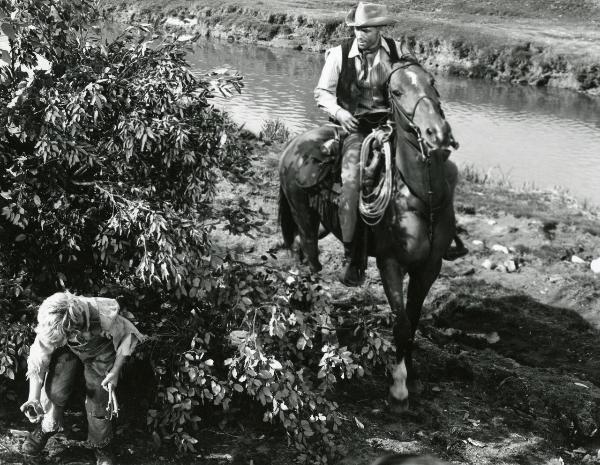 Scena del film "Un fiume di dollari" - Lizzani, Carlo, 1966 - Tom Hunter, a cavallo lungo un fiume, si volta verso sinistra per guardare un bambino dietro a una pianta che raccoglie sassi e legnetti.
