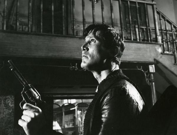 Scena del film "Un fiume di dollari" - Lizzani, Carlo, 1966 - Henry Silva di profilo con una pistola nella mano destra rivolge lo sguardo verso l'alto verso il soppalco sopra di lui.
