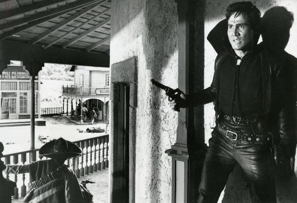 Scena del film "Un fiume di dollari" - Lizzani, Carlo, 1966 - Henry Silva nascosto e appoggiato a un muro con la pistola nella mano sinistra guarda davanti a sé. Alcuni attori non identificati che si spostano tra gli edifici.