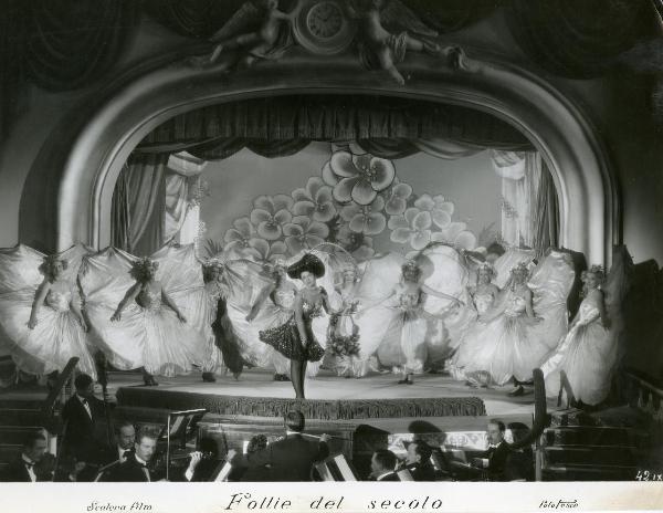 Scena del film "Follie del secolo" - Palermi, Amleto, 1939 - Scena di varietà parigino: un attrice non identificata in primo piano è circondata da altre attrici non identificate. Nella parte sottostante è presente l'orchestra.
