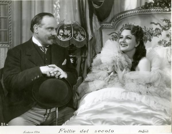 Scena del film "Follie del secolo" - Palermi, Amleto, 1939 - Paola Barbara a letto circondata da tulle guarda felice verso sinistra. Di fianco a lei è seduto un attore non identificato che la guarda appoggiando le mani sul bastone davanti a lui.

