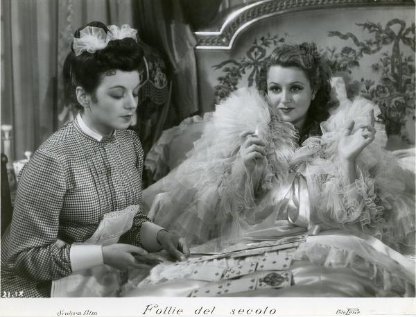 Scena del film "Follie del secolo" - Palermi, Amleto, 1939 - Clelia Matania, seduta di fianco al letto, posiziona le carte da gioco. Sul letto seduta e circondata da tulle, Paola Barbara.
