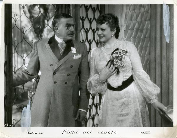 Scena del film "Follie del secolo" - Palermi, Amleto, 1939 - Paola Barbara in abito di pizzo si protende verso Armando Falconi, a sinistra, che la guarda con la mano destra sulla finestra già aperta.
