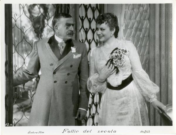 Scena del film "Follie del secolo" - Palermi, Amleto, 1939 - Paola Barbara in abito di pizzo si protende verso Armando Falconi, a sinistra, che la guarda con la mano destra sulla finestra già aperta.
