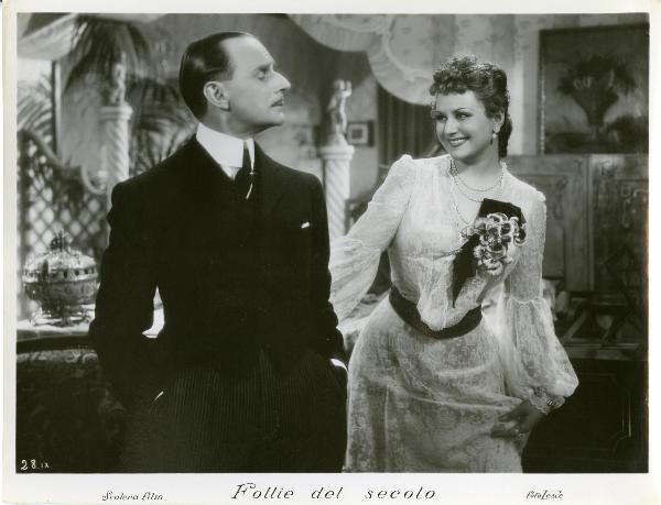 Scena del film "Follie del secolo" - Palermi, Amleto, 1939 - Paola Barbara, in veste di pizzo, protende la mano destra verso Sergio Tofano, davanti a lei che, con le mani nelle tasche dei pantaloni eleganti, la guarda.
