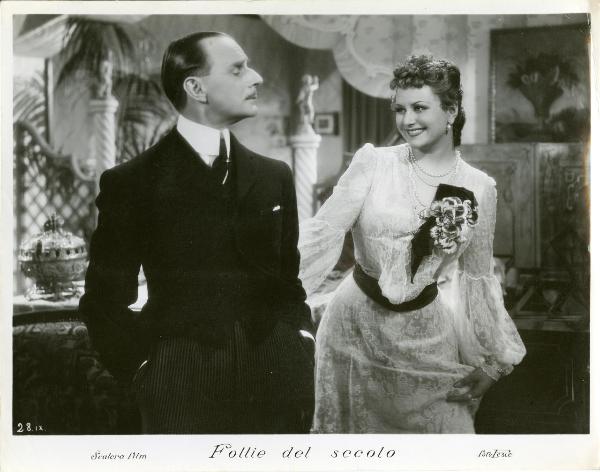 Scena del film "Follie del secolo" - Palermi, Amleto, 1939 - Paola Barbara, in veste di pizzo, protende la mano destra verso Sergio Tofano, davanti a lei che, con le mani nelle tasche dei pantaloni eleganti, la guarda.
