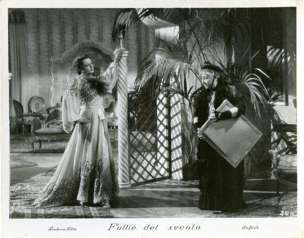 Scena del film "Follie del secolo" - Palermi, Amleto, 1939 - Paola Barbara appoggiata a un pilastro nella sua stanza, con la mano sinistra su un fianco, fissa una donna anziana con i vestiti sgualciti che tiene in mano un telaio rettangolare.