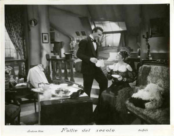 Scena del film "Follie del secolo" - Palermi, Amleto, 1939 - Paola Barbara seduta di fianco al divanetto con in mano una tazzina da té. Di fianco a lei sulla sinistra un attore non identificato che le porge una teiera.
