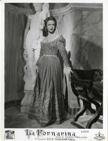 Scena del film "La fornarina" - Guazzoni, Enrico, 1942 - Anneliese Uhlig, in posa, si regge a una sedia sulla destra con la mano destra. In secondo piano è visibile una scultura bianca.

