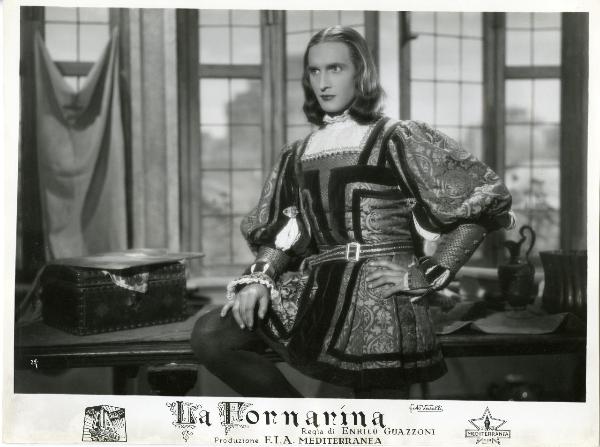 Scena del film "La fornarina" - Guazzoni, Enrico, 1942 - Walter Lazzaro, seduto con la gamba sinistra su un tavolo, fissa a sinistra con la mano destra su un ginocchio e a mano sinistra su un fianco.
