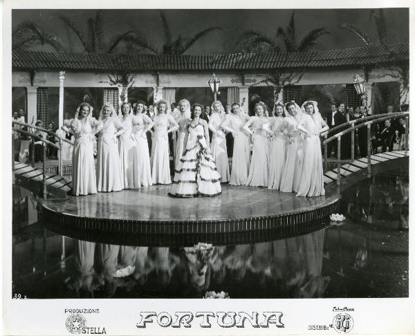 Scena del film "Fortuna" - Neufeld, Max, 1940 - Jone Salinas, al centro in abito elegante, è circondata da dodici attrici non identificate in posa con le mani sui fianchi.

