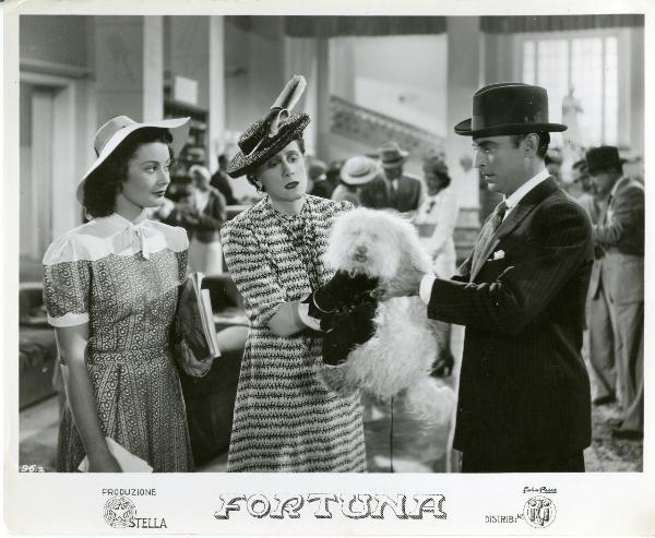 Scena del film "Fortuna" - Neufeld, Max, 1940 - Al centro, Julia Caba Alba tiene tra le mani un cane di piccola taglia e lo porge a Tony D'Algy che lo afferra con entrambe le mani. A sinistra, Maria Denis li osserva tenendo delle riviste.
