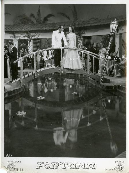 Scena del film "Fortuna" - Neufeld, Max, 1940 - Maria Denis, appoggiata a un ponte, guarda sorridendo Tony D'Algy sulla sinistra che la fissa appoggiandosi con entrambe le mani alla balaustra del ponte.

