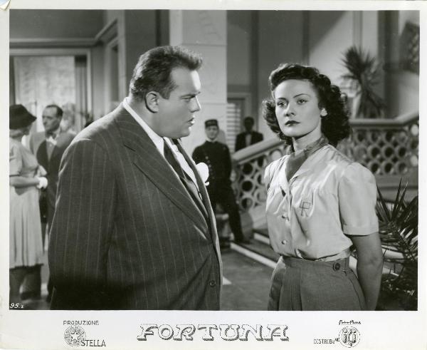 Scena del film "Fortuna" - Neufeld, Max, 1940 - Maria Denis, a destra, con le mani dietro la schiena guarda dritto davanti a sé mentre a sinistra Ugo Cesari le parla con un espressione minacciosa.
