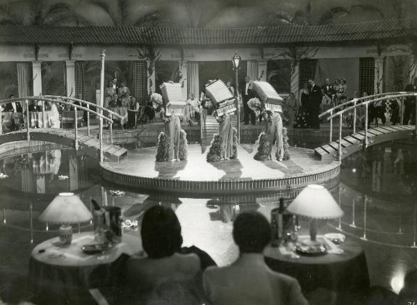 Scena del film "Fortuna" - Neufeld, Max, 1940 - Tre attrici non identificate su un palco, al centro, suonano delle fisarmoniche. Intorno a loro, attrici e attori non identificati le osservano.
