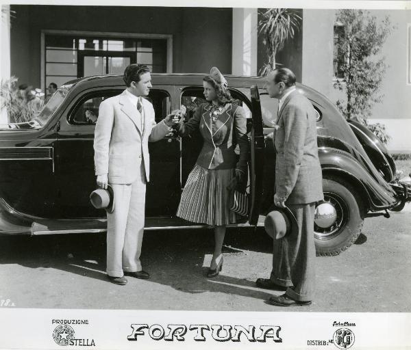 Scena del film "Fortuna" - Neufeld, Max, 1940 - Jone Salinas scende dall'auto aiutata da Luis Prendes. A destra, un attore non identificato tiene aperta la portiera dell'auto mentre li osserva.
