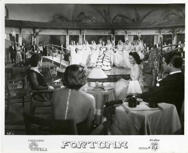 Scena del film "Fortuna" - Neufeld, Max, 1940 - Al centro del palco, Jone Salinas. Dietro di lei, dodici attrici non identificate con la mano destra che tiene un lembo della lunga gonna bianca. In primo piano, Maria Denis seduta.
